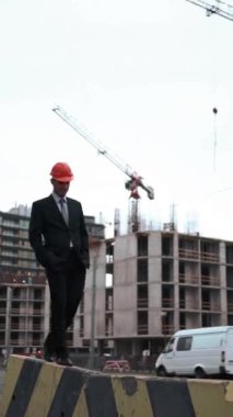 4k, UND. Takım elbiseli ve kask giyen genç bir mimar kirişin üzerinde vinç ve arka planda yeni inşa edilmiş bir binayla yürüyor. Teal ve turuncu stil. Statik Geniş Çekim Dikey Çekim.