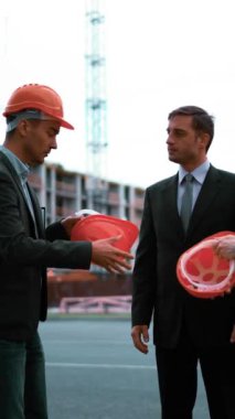 4 bin. UltraHD. Genç mühendis, takım elbiseli iş adamlarına kask takıyor: benimle çalışmaya gel. Binanın orada vinç ve kirişler var. Teal ve turuncu statik orta çekim. Dikey Çekim.