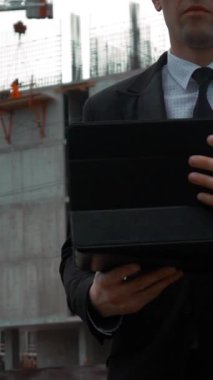 Siyah takım elbiseli genç ve başarılı müşteri işadamı, arka planda vinç ve kirişlerle yeni inşaat alanlarının yakınındaki dijital bloknotları kontrol edip kullanıyor. Orta buharlı iğne. Dikey Çekim.