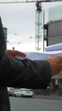 Ağır çekim. Proje yöneticisi, arka planında vinç ve kirişler olan yeni inşa edilmiş bir bina ile pano inşaat planını kontrol ediyor. Metin kağıdının detaylarına kadar. Dikey Çekim.