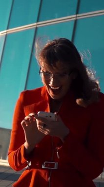 Ağır çekim. Kırmızı takım elbiseli ve gözlüklü seksi kadın akıllı telefonunun mesaj okumasını izliyor, arka planda ışıltılı modern iş merkeziyle seviniyor. Orta atış. Dikey