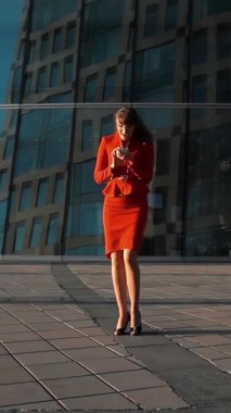Kırmızı takım elbiseli neşeli seksi iş kadını ve iş bölgesindeki gökdelenin yanındaki anlaşmayı kazanma sevinci içinde akıllı telefon, 240 fps 'te yavaş çekim, sabit kamera çekimi. Dikey Çekim.