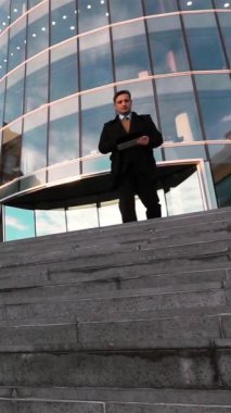 Ağır çekim: İş adamı yürüyor ve kentsel çevrede dijital ped kullanıyor, cam iş merkezi bg. Teal ve Turuncu. Geniş açı. Yüksek hızlı kamera. Dikey Çekim.