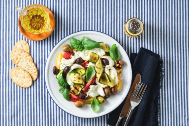 Akdeniz salatası, mozarella, zeytin, fesleğen ve ızgara sebze ile mavi çizgili keten masa örtüsü.