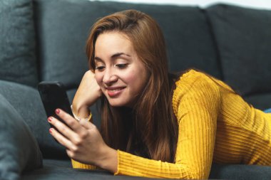 Kahverengi saçlı, sarı gömlekli, kot giyen, koltukta oturan, cep telefonu kullanan bir kadın..