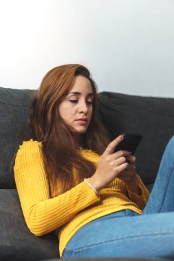 Kahverengi saçlı, sarı gömlekli, kot giyen, koltukta oturan, cep telefonu kullanan bir kadın..