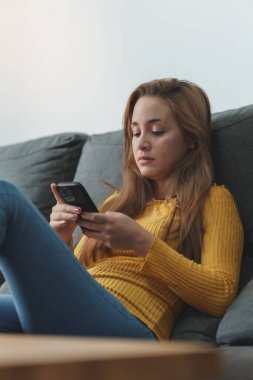 Kahverengi saçlı, sarı gömlekli, kot giyen, koltukta oturan, cep telefonu kullanan bir kadın..