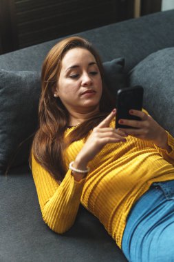 Kahverengi saçlı, sarı gömlekli, kot giyen, koltukta oturan, cep telefonu kullanan bir kadın..