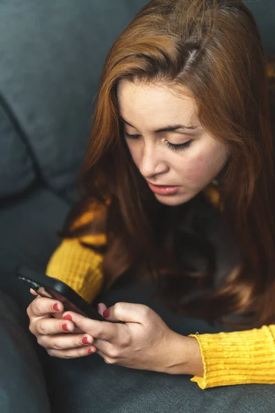 Kahverengi saçlı, sarı gömlekli, kot giyen, koltukta oturan, cep telefonu kullanan bir kadın..