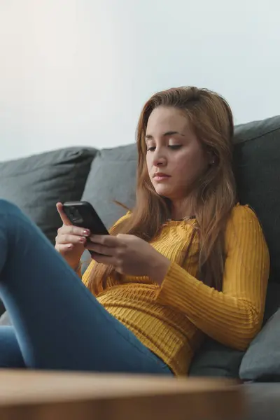 Kahverengi saçlı, sarı gömlekli, kot giyen, koltukta oturan, cep telefonu kullanan bir kadın..