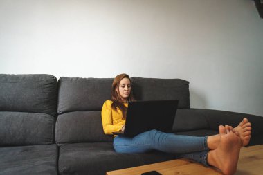 Sarı ceketli ve kot pantolonlu bir kadın koltukta bilgisayar kullanıyor.