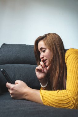 Sarı ceketli ve kot pantolonlu bir kadın koltukta cep telefonu kullanıyor.