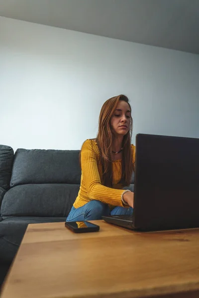 Sarı ceketli ve kot pantolonlu bir kadın koltukta bilgisayar kullanıyor.