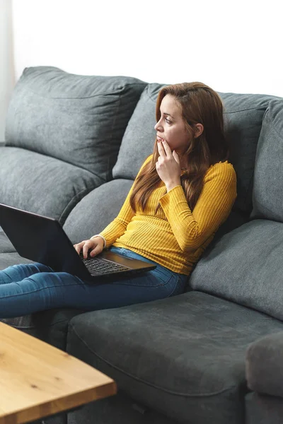 Sarı ceketli ve kot pantolonlu bir kadın koltukta bilgisayar kullanıyor.