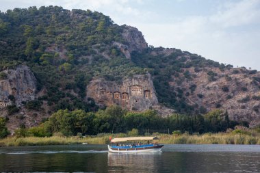 Dalyan - Mugla - Türkiye, 22 Ekim 2024, Dalyan, Mugla, Türkiye 'deki Kaunos antik kentinin kaya kesimli tapınak mezarları. Feribot ve arka planda oyulmuş mezarlarla güzel Dalyan nehir manzarası.