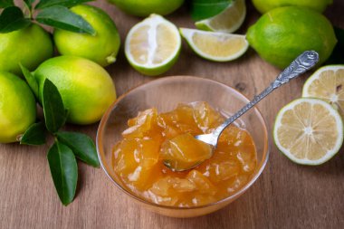 Beyaz ahşap bir masada limon reçeli kavanozu. Masada limon dilimleri ve yeşil yapraklar var..