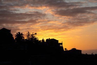 Puerto de la Cruz, Tenerife 'de gün batımında canlı turuncu ve sarı gökyüzü ile bir yamaçta ev ve palmiye ağaçları silueti..