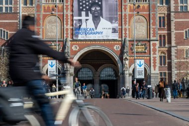 Amsterdam, Hollanda - 20 Mart 2025 Kargo motosikletli bisikletçi Amerikan Fotoğraf Sergisi bayrağı altında Rijksmuseum 'dan geçiyor