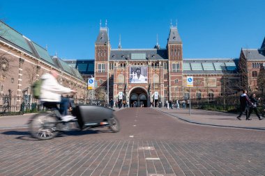 Amsterdam, Hollanda - 20 Mart 2025 Kargo motosikletli bisikletçi Amerikan Fotoğraf Sergisi bayrağı altında Rijksmuseum 'dan geçiyor