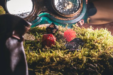 Kırmızı d20, turuncu d10 ve siyah zar Steampunk gözlükleri ve fantezi paralarının yanındaki yosunların üzerinde dinleniyor..