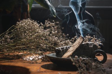 Palo Santo ve adaçayı lekesi görünür dumanla yanıyor, ahşap masada kurumuş çiçeklerle çevrili.