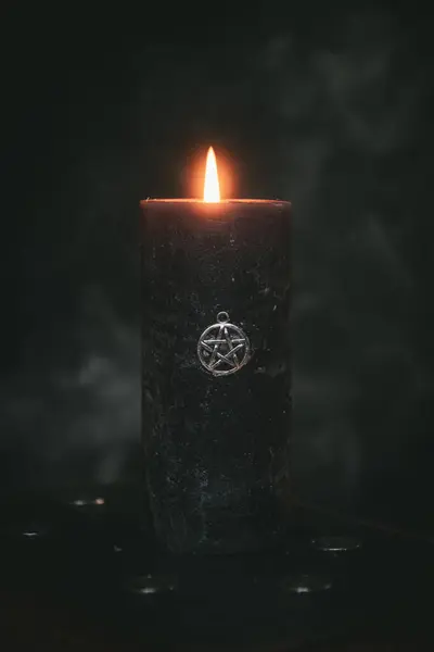 Gümüş bir pentagram kolyeli siyah mum, karanlıkta parlayan mistik bir ortam ve arka planda hafif bir duman.