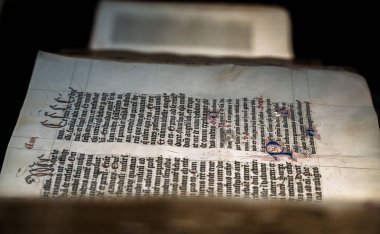 Gotik yazı, dekoratif baş harfleri ve elle boyanmış süslü detayların yer aldığı aydınlatılmış bir ortaçağ el yazması sayfasının yakın çekimi. Ortaçağ kaligrafi ve işçiliğini gösteren tarihi belge.