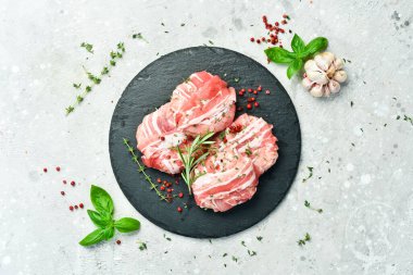 Biberiye ve kekik ile birlikte siyah bir tabakta servis edilen domuz pastırmalı çiğ pirzola. Gri bir arka planda. Üst görünüm.