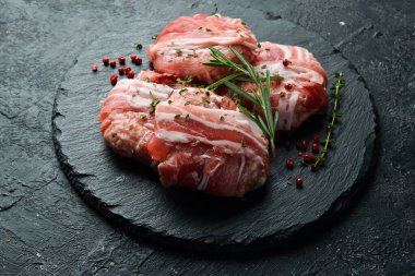 Domuz pastırmasına sarılı çiğ pirzola, siyah bir tabakta biberiye ve kekik ile servis edilir. Taş zeminde. Üst görünüm.