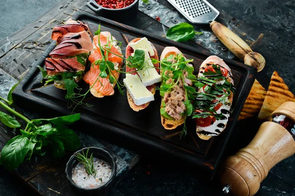 stock image sandwich Set bruschetta with tuna, cheese, prosciutto and tomatoes on a dark wooden board. On a black stone background.