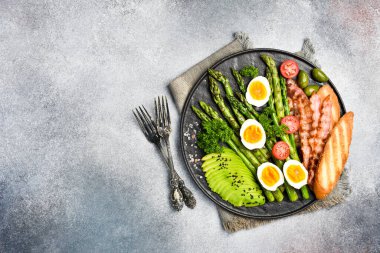 Kahvaltı: kuşkonmaz, avokado, tavuk yumurtası ve pastırma. Taş bir tabakta. Gri beton bir tuvalde.