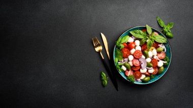 Bir kasede Caprese salatası: domates, mozzarella peyniri, fesleğen ve zeytin. Siyah taştan bir tuvalde. Minimalizm tarzı. Üst görünüm.