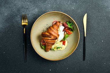 Kahvaltıda. Pastırmalı kruvasan, haşlanmış yumurta, marul ve domates. Siyah taştan bir arka planda. Metin için boş alan.