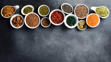 Süper yiyecekler, tahıllar, fındıklar ve kurutulmuş meyveler, spirulina ve goji seti beyaz seramik kaselerde. Gri beton arka planda. Üst Manzara. Metin için boş alan.