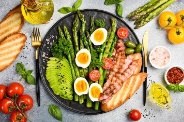 Besleyici Kahvaltı: yeşil kuşkonmaz, avokado, tavuk yumurtası ve pastırma. Taş bir tabakta.