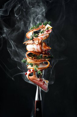 Slices of smoked bacon on a metal fork. On a black background, close-up. clipart