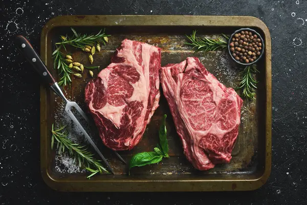 Metal bir tepside baharat ve otlarla birlikte iki çiğ mermer biftek. Pişirmeye hazır. Kırsal biçim.