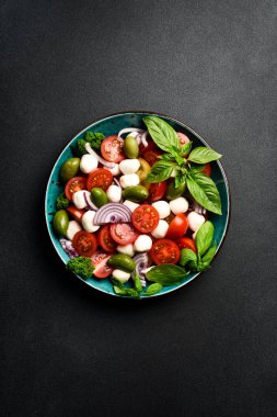Bir kasede Caprese salatası: domates, mozzarella peyniri, fesleğen ve zeytin. Siyah taştan bir tuvalde. Minimalizm tarzı. Üst görünüm.