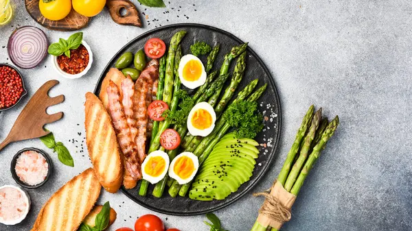 Besleyici Kahvaltı: yeşil kuşkonmaz, avokado, tavuk yumurtası ve pastırma. Taş bir tabakta.