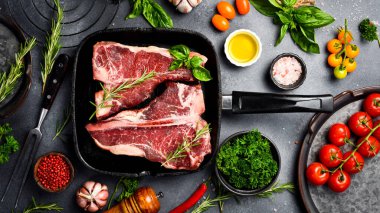 Tavada çiğ T-Kemik Bifteği. Gri bir masada biftek, baharat, baharat, tereyağı ve tuz pişirmek..