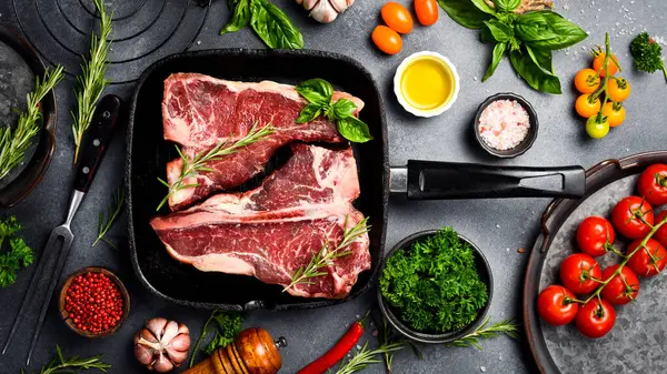 Tavada çiğ T-Kemik Bifteği. Gri bir masada biftek, baharat, baharat, tereyağı ve tuz pişirmek..