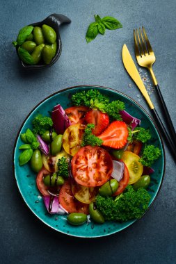 Farklı türde domates, soğan ve zeytinlerden yapılmış maydanoz ve fesleğenle süslenmiş salata kasesinin üst görüntüsü. Siyah çimento bir masada..