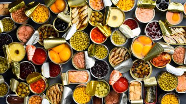 Arka planda turşulu ve konserve yiyecekler var. Konserve sebzeler, fasulyeler, balıklar, deniz ürünleri ve meyvelerle metal kutular. Üst görünüm.