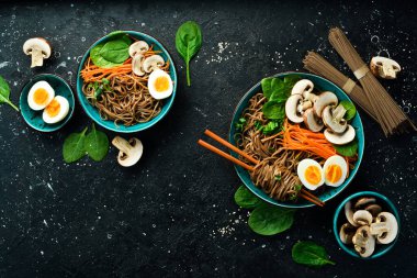 Bir kase mantarlı, havuçlu, soğanlı ve yumurtalı Japon eriştesi. Taş zeminde. Üst görünüm.