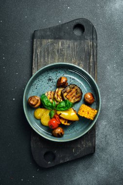 Bir tabak ev yapımı ızgara sebze: patlıcan, mısır, kırmızı biber ve domates. Siyah taştan bir arka planda. Metin için boş alan.