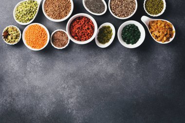 Süper yiyecekler, tahıllar, fındıklar ve kurutulmuş meyveler, spirulina ve goji seti beyaz seramik kaselerde. Gri beton arka planda. Üst Manzara. Metin için boş alan.