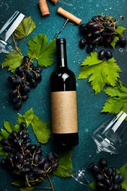 Kırmızı şarap şişeleri, üzümler, bardaklar, tirbuşon ve siyah desenli masa mantarları. Üst görünüm.