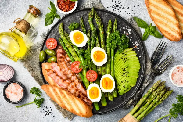 Besleyici Kahvaltı: yeşil kuşkonmaz, avokado, tavuk yumurtası ve pastırma. Taş bir tabakta.