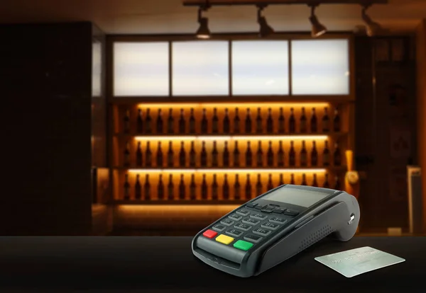 credit card reader pos machine and a credit card are on wooden table with background of beer bottle shelf in an empty nice bar in evening during cleaning process before opening to serve customers