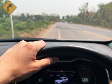 driving on a rural road with a visible curve warning sign, showing a close-up of a left hand holding the steering wheel inside a car, focusing on safe travel and road journey atmosphere dashboard view